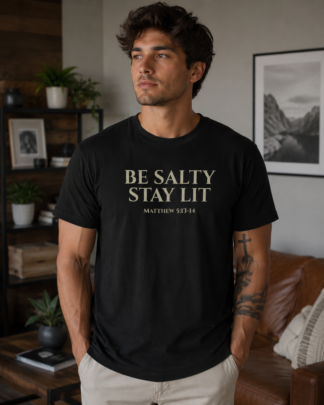 Young man wearing a black Christian “Be Salty Stay Lit Matthew 5:13-14” t-shirt while standing in a cozy modern living room with warm neutral decor and soft natural lighting