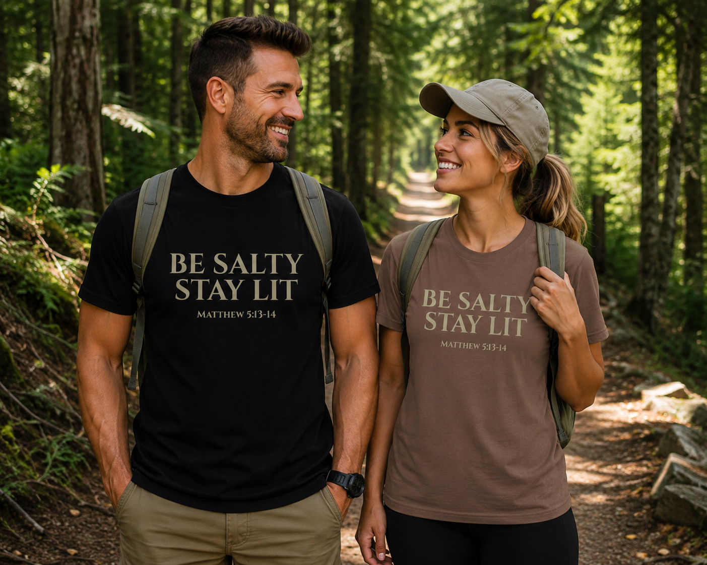 Man wearing a black “Be Salty Stay Lit Matthew 5:13-14” Christian t-shirt and woman wearing an espresso-colored version walking together on a wooded hiking trail with backpacks and a warm outdoor lifestyle aesthetic