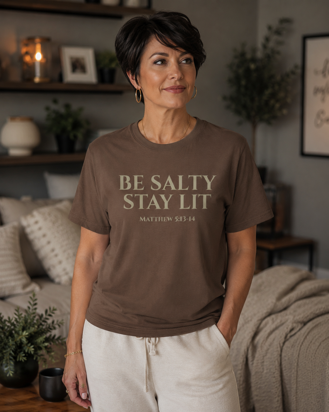 Middle-aged woman with short dark hair wearing an espresso-colored Christian “Be Salty Stay Lit Matthew 5:13-14” t-shirt while standing in a cozy modern living room with warm neutral decor and soft ambient lighting
