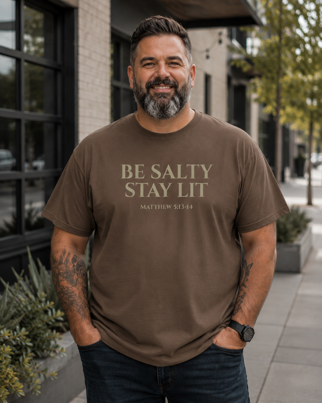 Plus-size middle-aged man with a beard wearing an espresso-colored Christian “Be Salty Stay Lit Matthew 5:13-14” t-shirt while standing outdoors in a modern city setting with soft natural lighting and an urban backdrop