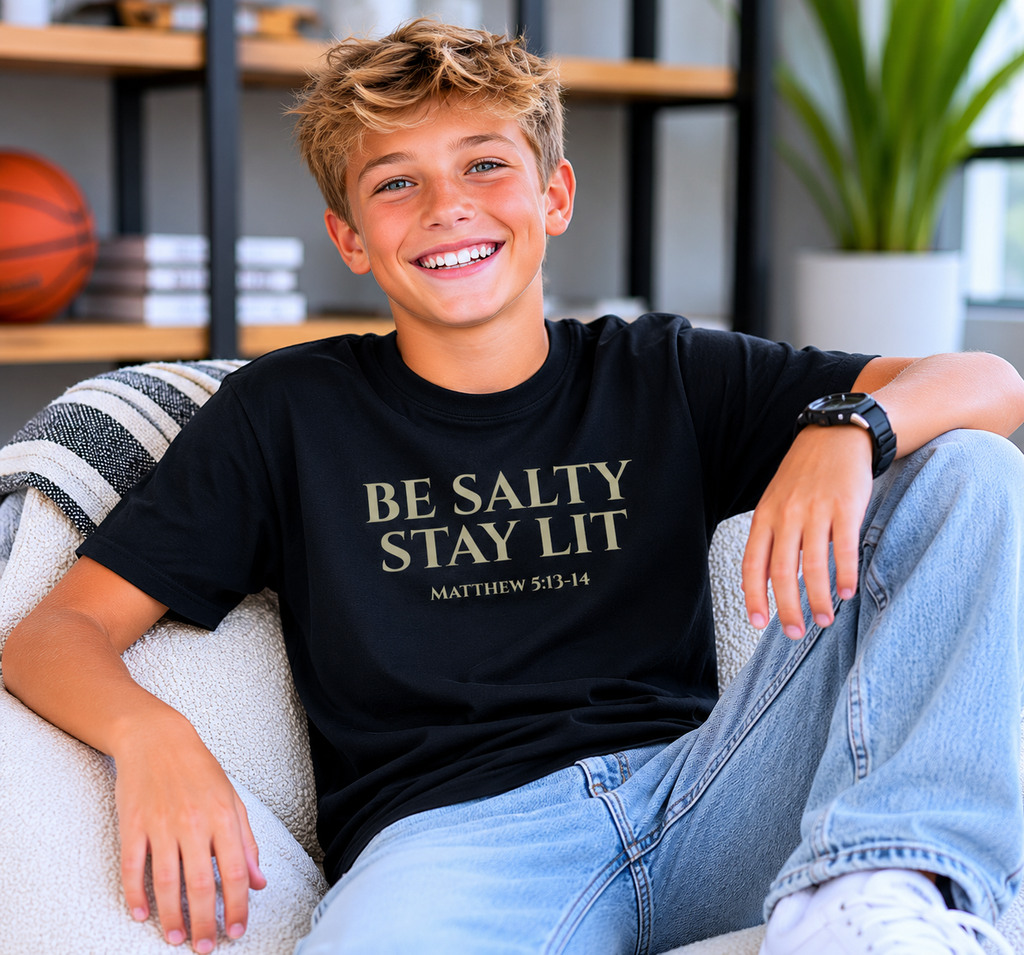 Tween boy with short blonde hair wearing the black “Be Salty Stay Lit” youth Christian t-shirt in a modern indoor gaming-inspired room with a fun, youthful vibe.