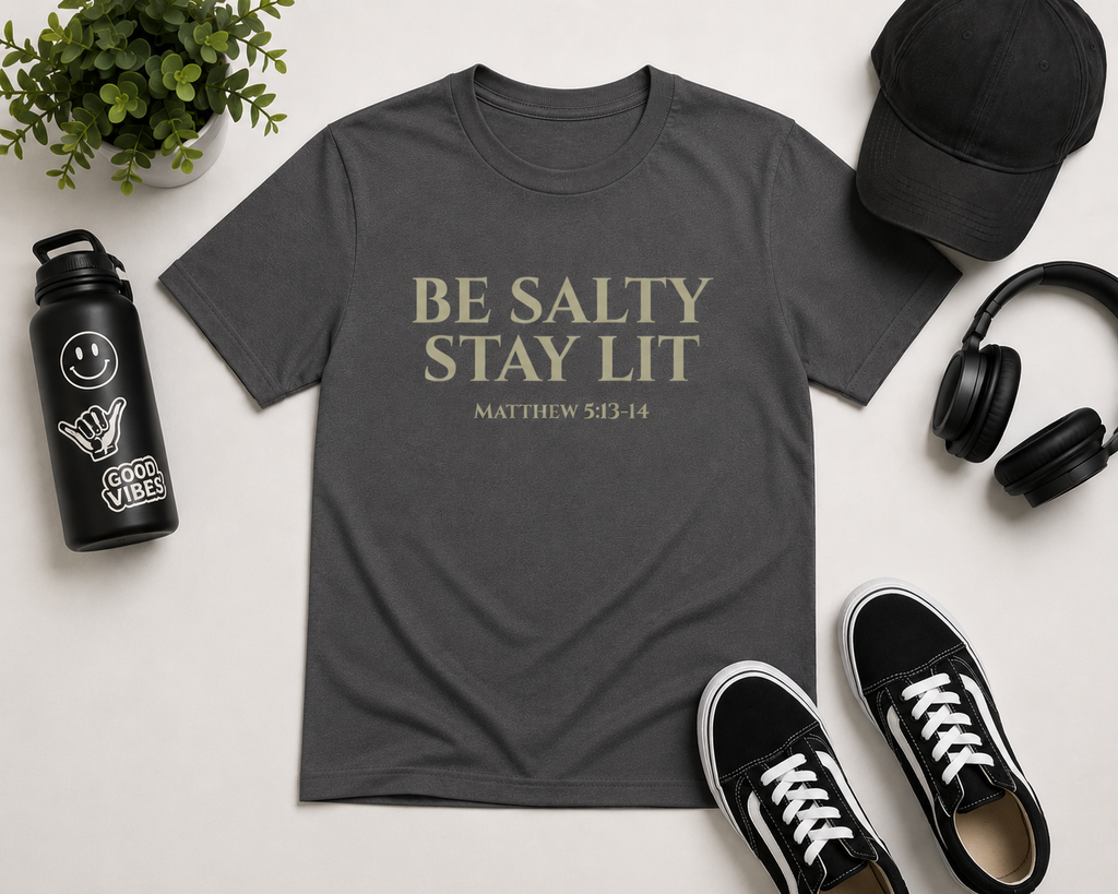 Flat lay mockup of the pepper gray “Be Salty Stay Lit” youth Christian t-shirt styled with black sneakers, headphones, a black cap, water bottle, and minimal modern accessories on a clean neutral background.
