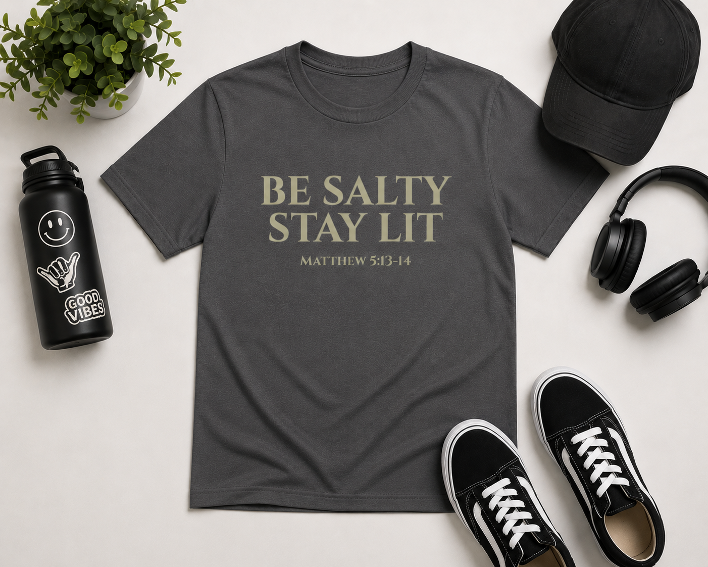 Flat lay mockup of the pepper gray “Be Salty Stay Lit” youth Christian t-shirt styled with black sneakers, headphones, a black cap, water bottle, and minimal modern accessories on a clean neutral background.