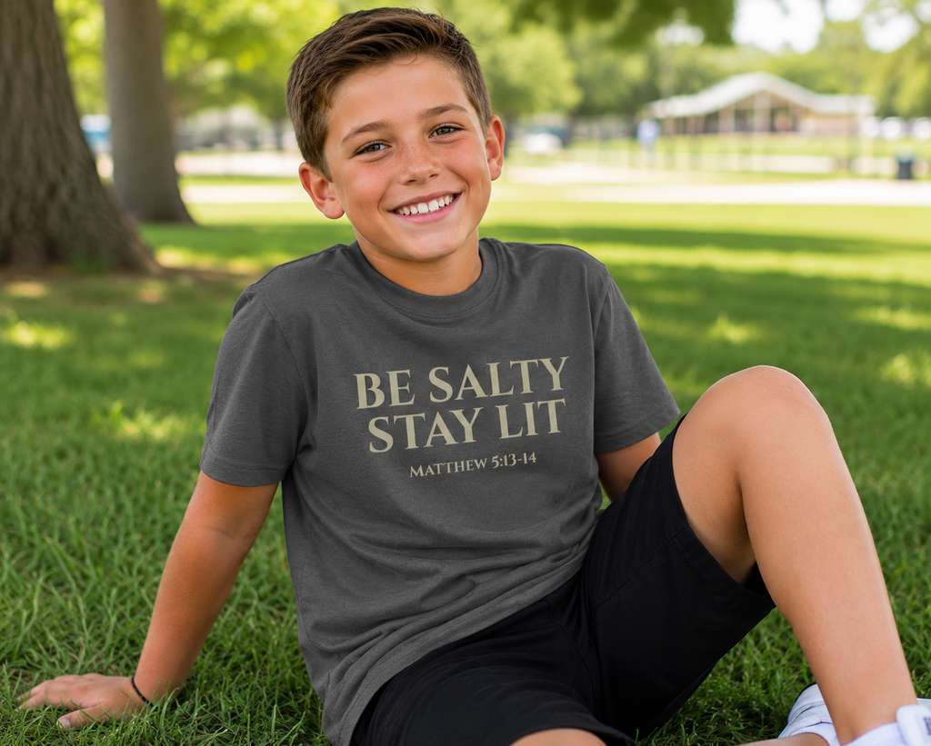 Young boy with short brown hair wearing the pepper gray “Be Salty Stay Lit” youth Christian t-shirt while relaxing in a grassy park on a sunny day with a fun, modern vibe.
