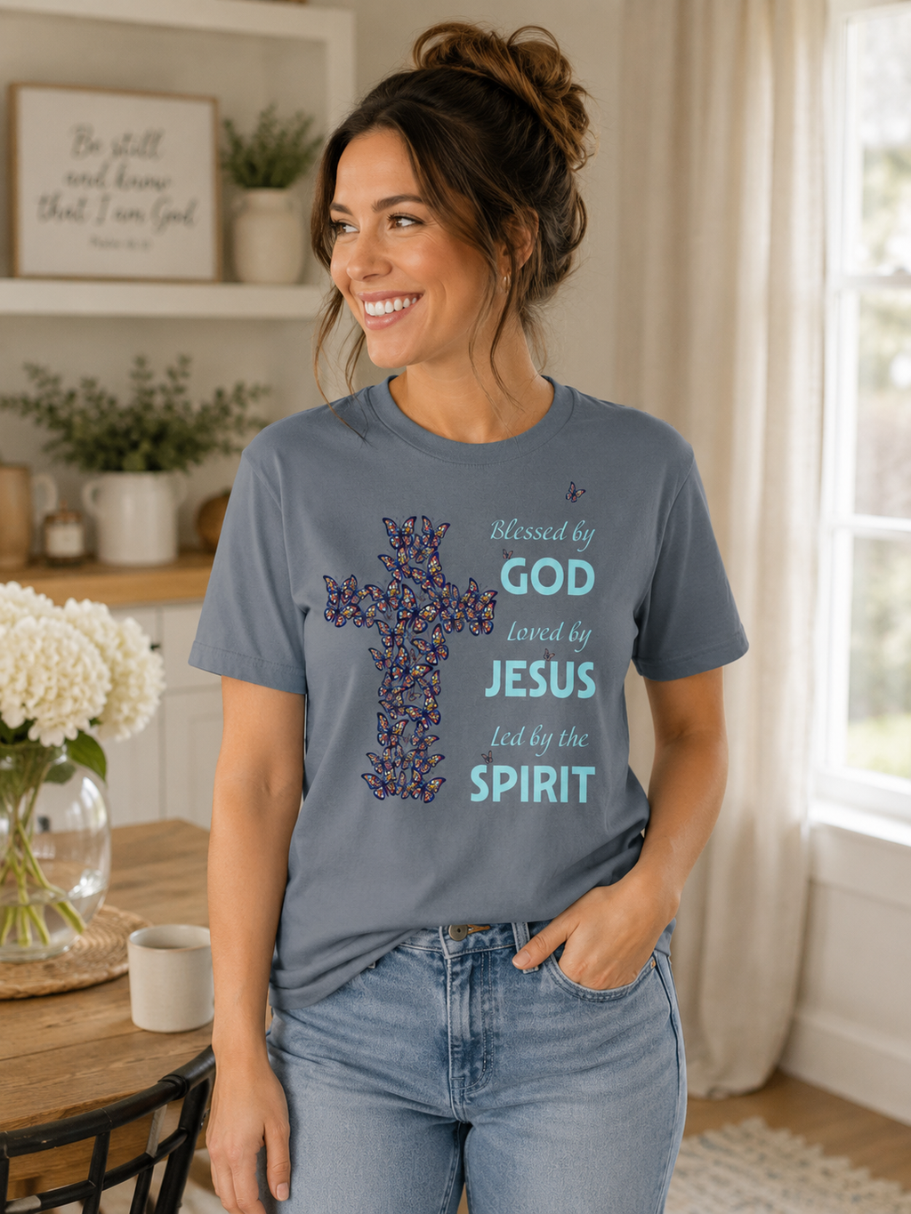 Woman wearing pepper blue Christian butterfly cross t-shirt with “Blessed by God, Loved by Jesus, Led by the Spirit” message in a cozy neutral-toned home with soft natural lighting