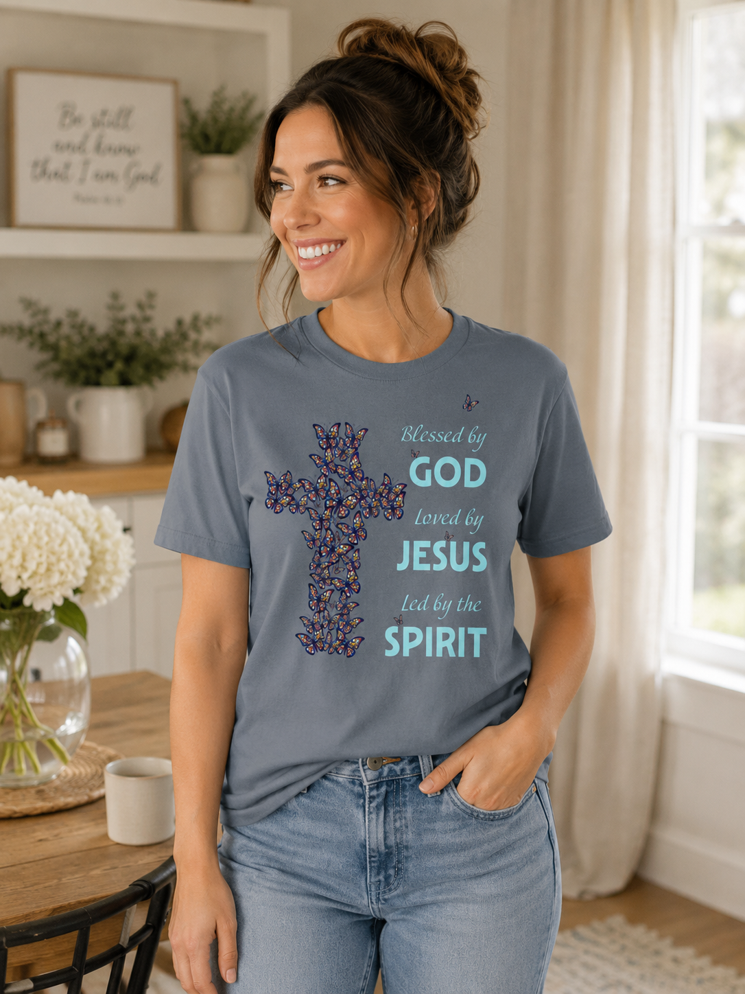 Woman wearing pepper blue Christian butterfly cross t-shirt with “Blessed by God, Loved by Jesus, Led by the Spirit” message in a cozy neutral-toned home with soft natural lighting