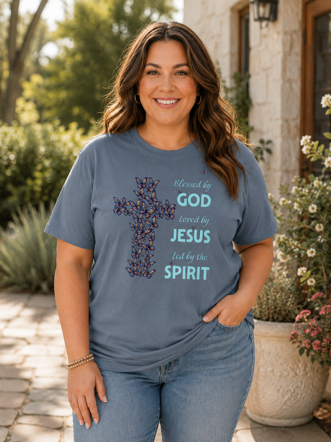 Plus size woman wearing pepper blue Christian butterfly cross t-shirt with inspirational faith message while standing outdoors in soft sunlight near greenery and stone architecture
