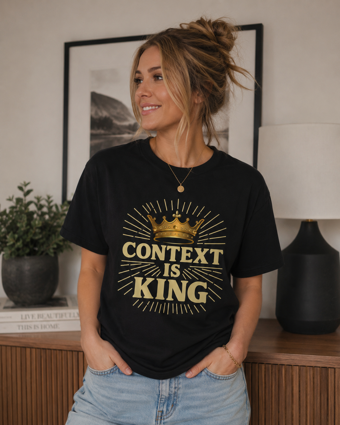 Woman in her 30s with dirty blonde hair in a messy updo wearing a black “Context Is King” Christian graphic t-shirt while standing in a cozy modern living room with warm neutral decor and soft lighting