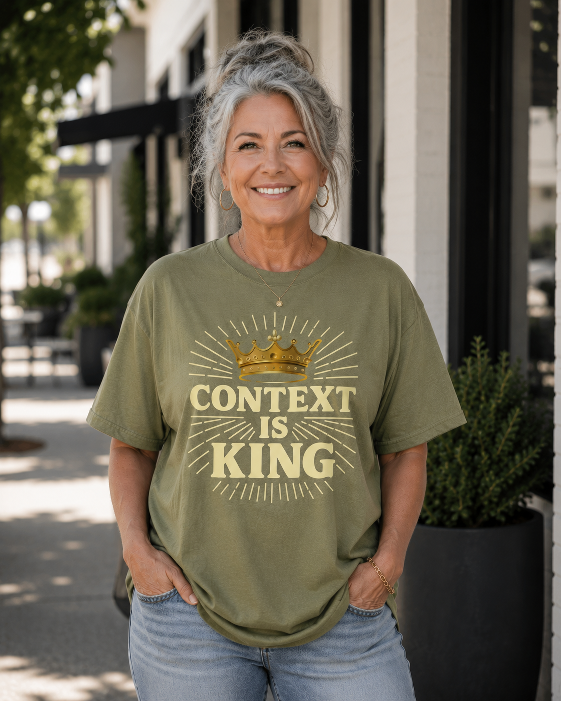 Older woman with salt-and-pepper hair wearing a hemp-colored “Context Is King” Christian graphic t-shirt while standing outdoors on a modern city sidewalk with natural lighting and a relaxed confident smile