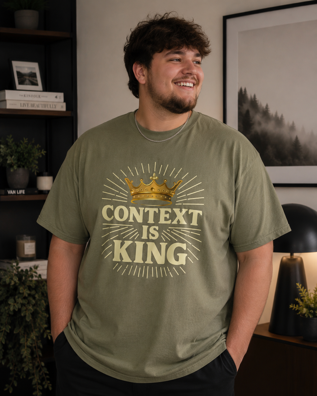 Young plus-size man wearing a hemp-colored “Context Is King” Christian graphic t-shirt while standing in a cozy modern living room with warm lighting, contemporary decor, and a relaxed casual style
