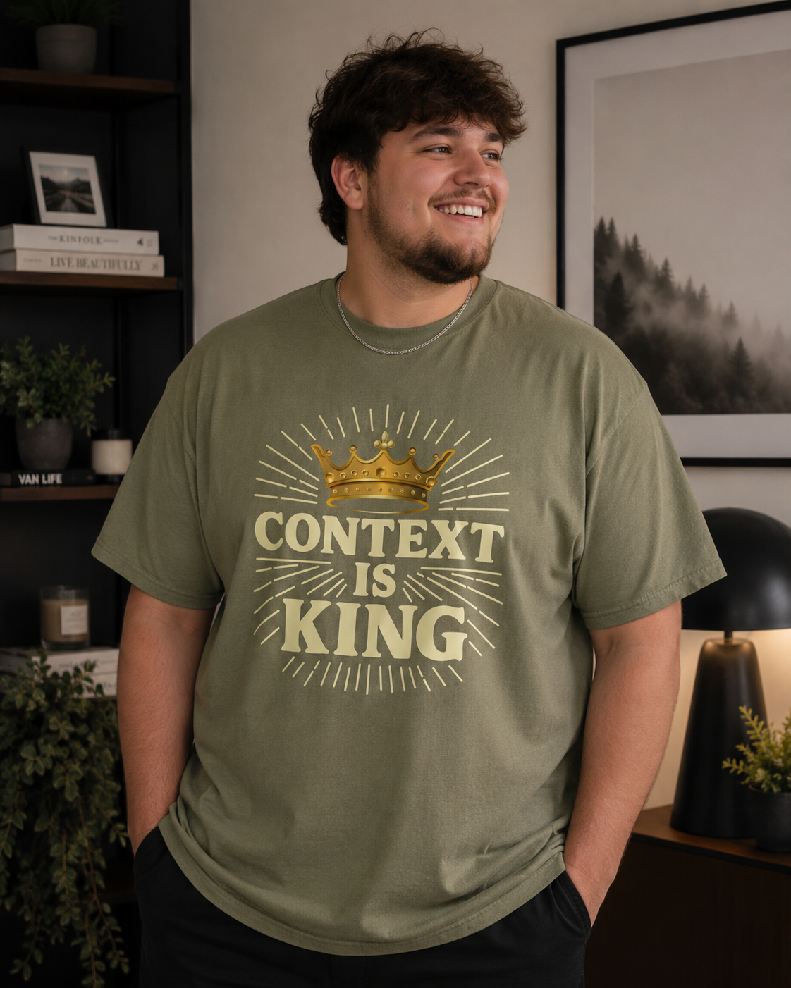 Young plus-size man wearing a hemp-colored “Context Is King” Christian graphic t-shirt while standing in a cozy modern living room with warm lighting, contemporary decor, and a relaxed casual style