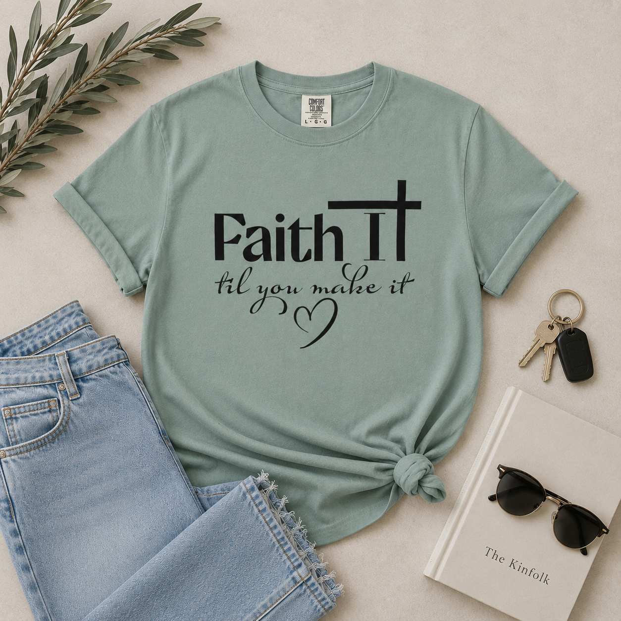 Bay blue “Faith It Til You Make It” Christian t-shirt flat lay with light denim, sunglasses, keys, and neutral modern styling on a beige background