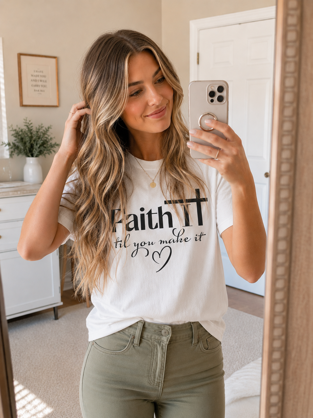Woman with long wavy blonde hair wearing white “Faith It Til You Make It” Christian t-shirt with olive green pants while taking a mirror selfie in a softly lit neutral bedroom