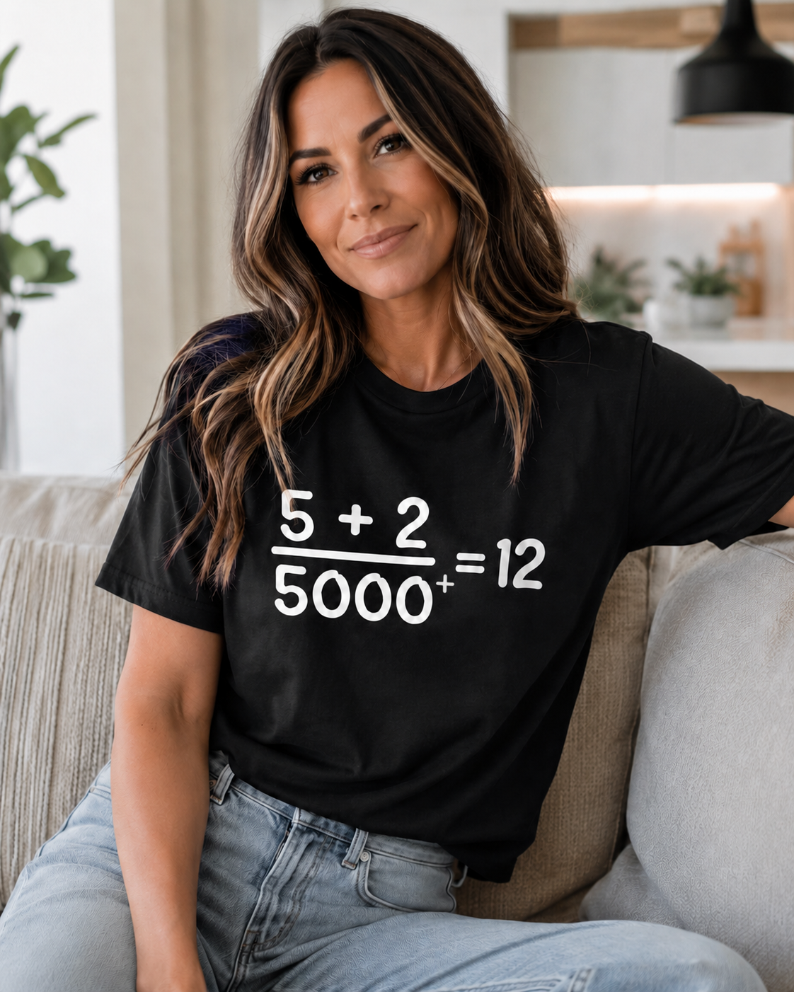 Woman with long brown hair and blonde highlights wearing a black Comfort Colors Christian math graphic t-shirt indoors in a modern neutral home setting featuring the “5 + 2 / 5000+ = 12” design.