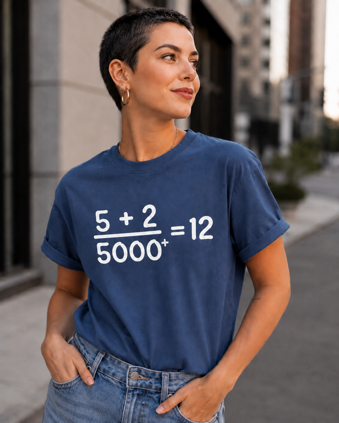 Young woman with very short dark hair wearing a china blue Comfort Colors Christian math graphic t-shirt in a modern city setting featuring the “5 + 2 / 5000+ = 12” miracle design.