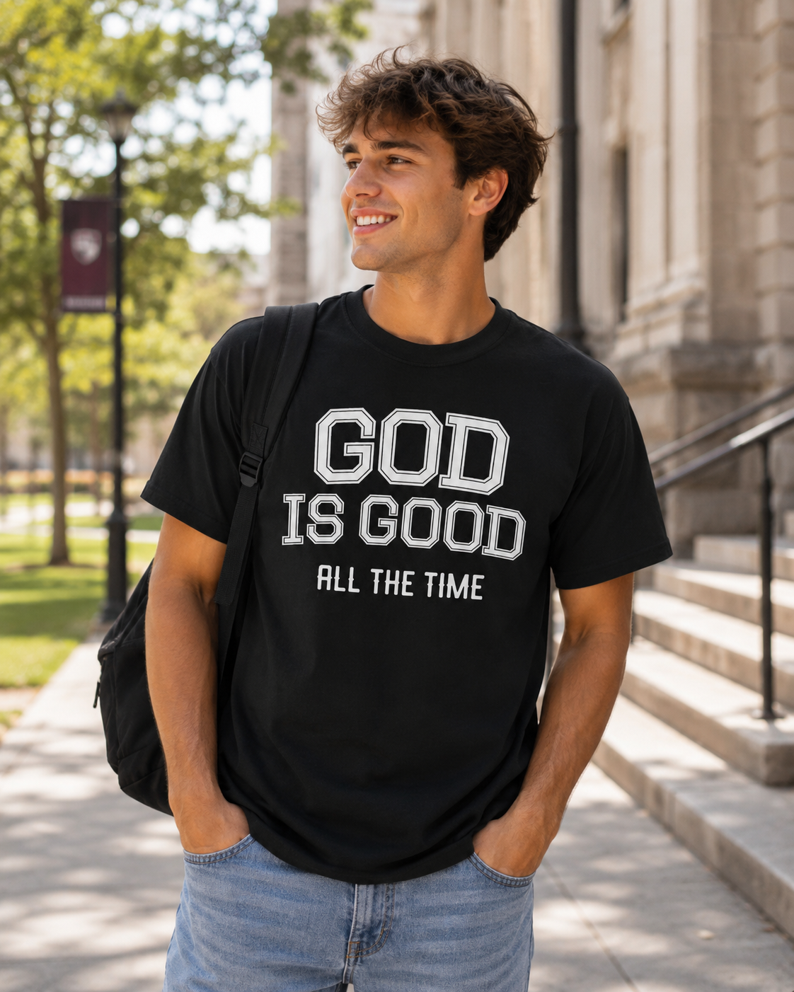 Young man wearing black Christian “God Is Good All The Time” t-shirt while walking on a sunny college campus with a backpack and casual modern styling