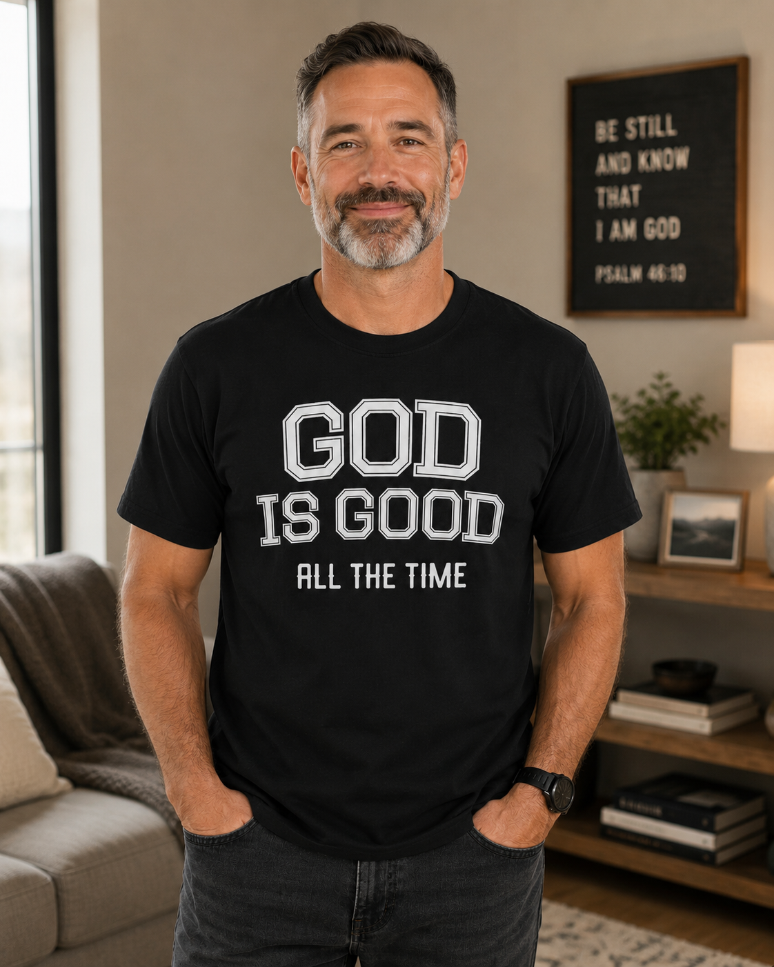 Older man wearing black Christian “God Is Good All The Time” t-shirt while standing in a cozy modern living room with warm neutral decor and soft lighting