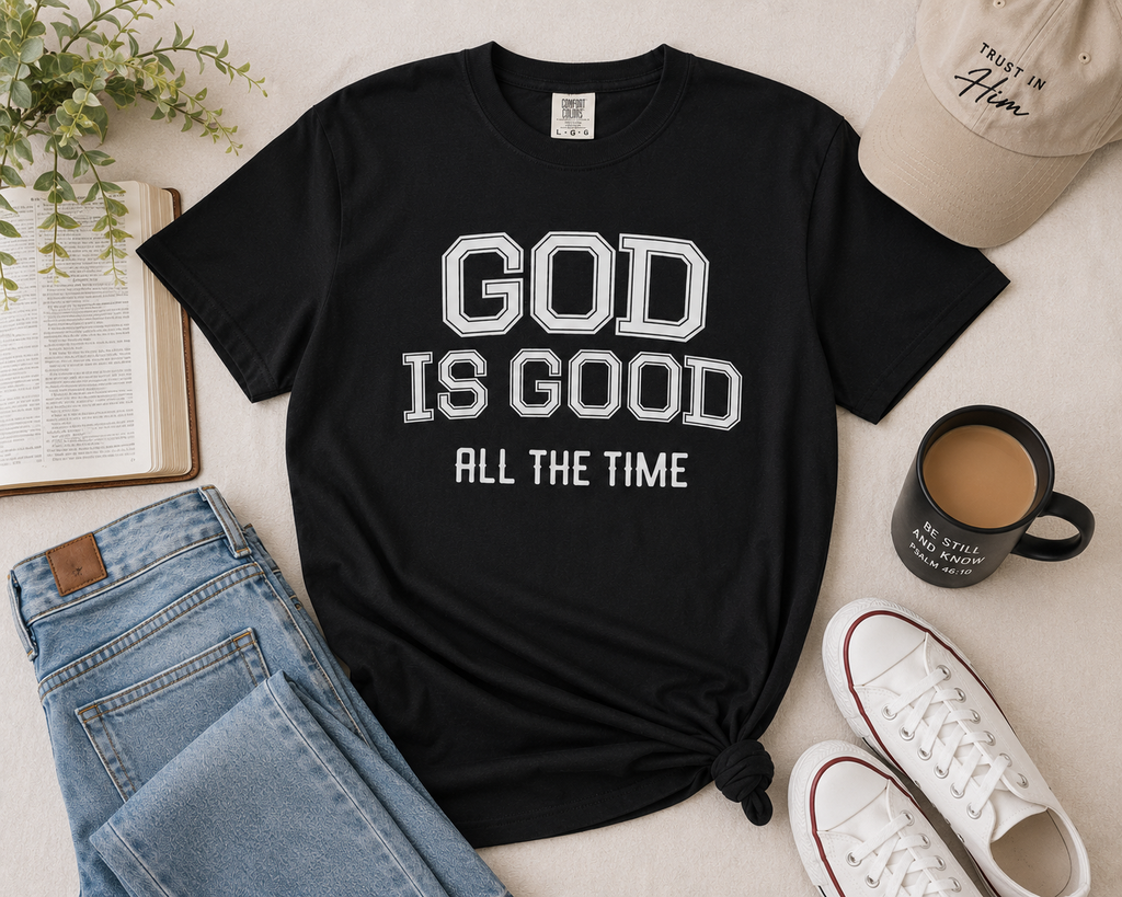 Black Christian “God Is Good All The Time” t-shirt flat lay styled with denim, neutral sneakers, a Bible, coffee mug, and modern faith-inspired accessories on a light neutral background