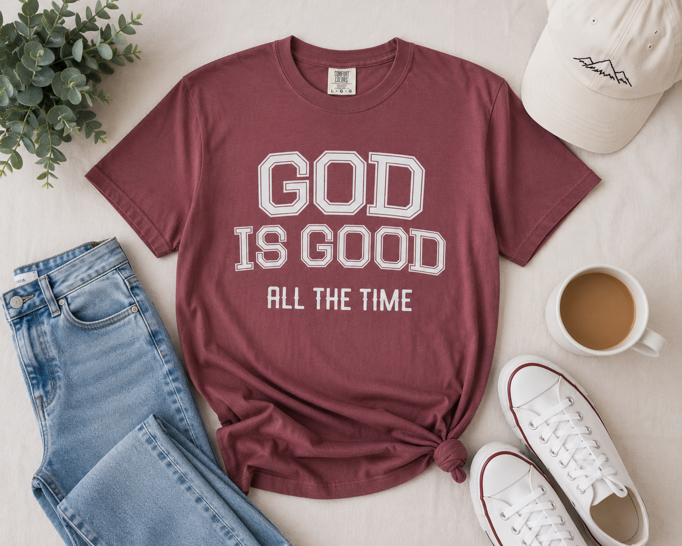 Brick-colored Christian “God Is Good All The Time” t-shirt flat lay styled with light denim, neutral sneakers, greenery, and modern casual accessories on a soft beige background