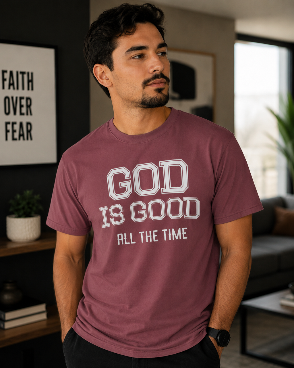 Man with a goatee wearing brick-colored Christian t-shirt with “God Is Good All The Time” text while standing in a modern cozy living room with warm neutral decor and soft natural lighting