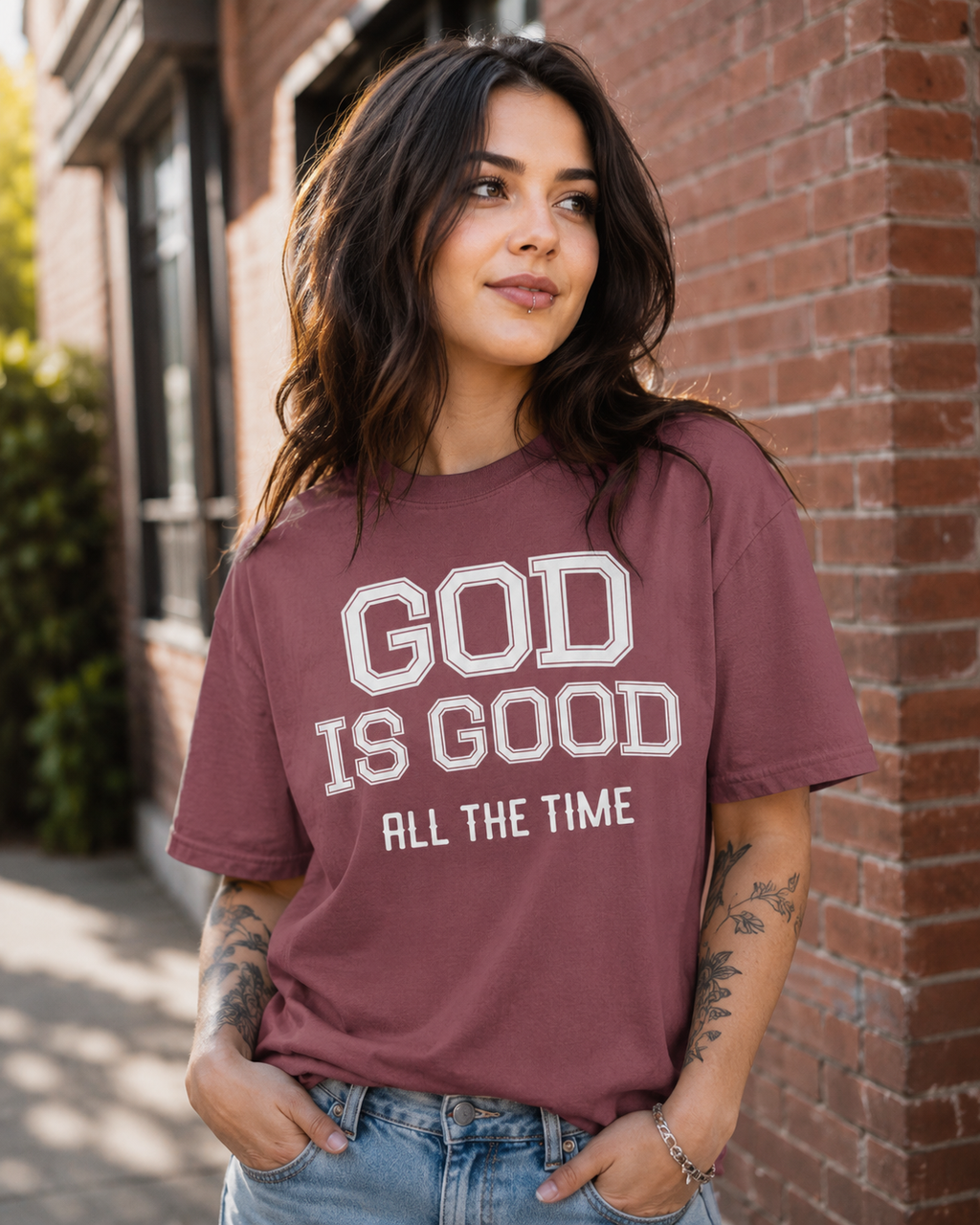 Woman with dark hair and a lip piercing wearing brick-colored Christian t-shirt with “God Is Good All The Time” text while standing outdoors beside a brick wall in soft natural sunlight