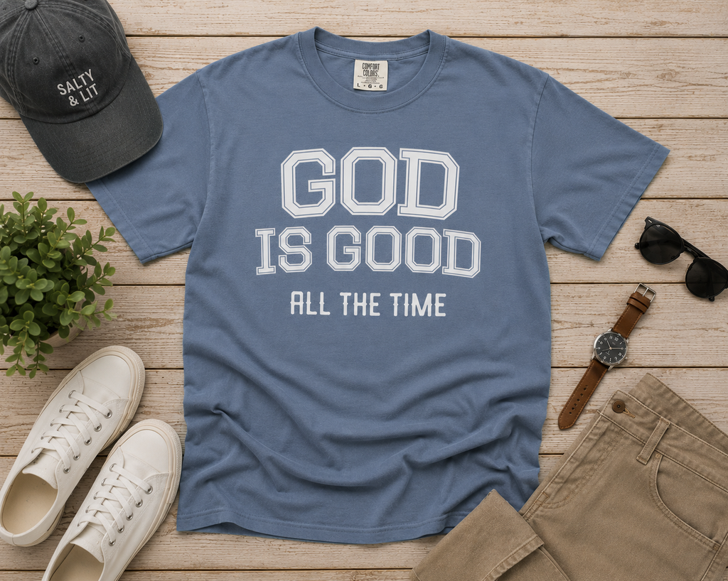 Flat lay of the God Is Good All The Time Christian T-Shirt in Blue Jean Comfort Colors 1717, styled with neutral men's accessories including white sneakers, khaki pants, a charcoal cap, sunglasses, and a leather watch on a light wood background.
