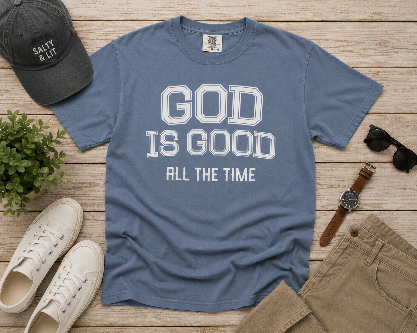 Flat lay of the God Is Good All The Time Christian T-Shirt in Blue Jean Comfort Colors 1717, styled with neutral men's accessories including white sneakers, khaki pants, a charcoal cap, sunglasses, and a leather watch on a light wood background.