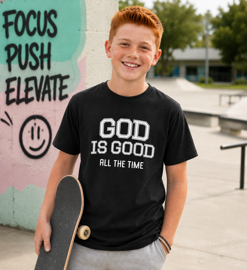 Tween boy with short red hair wearing the black “God Is Good All The Time” youth Christian t-shirt at an outdoor skatepark, holding a skateboard with a modern fun youthful vibe.
