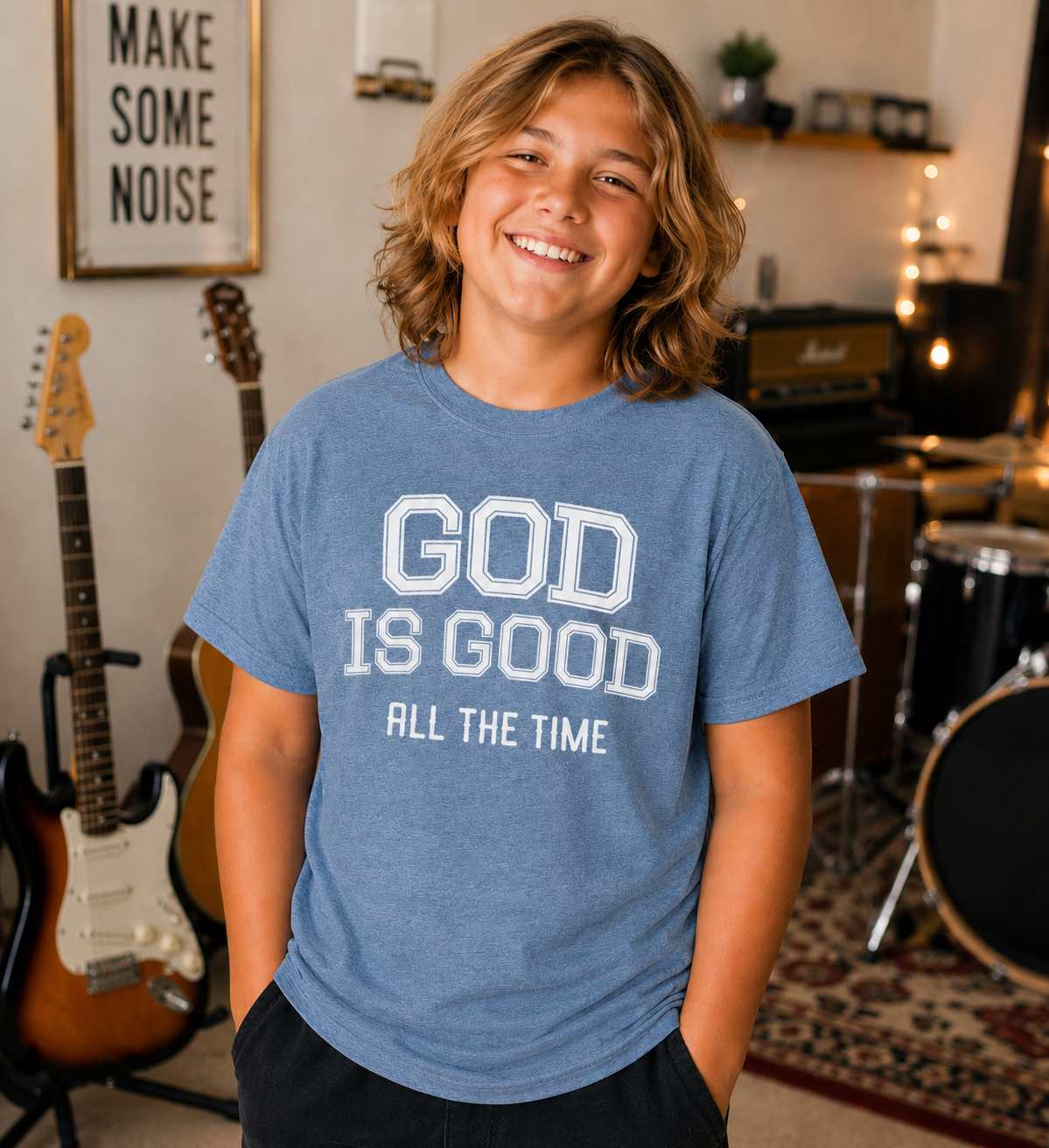 Tween boy with shoulder-length dirty blonde hair wearing the Blue Jean “God Is Good All The Time” youth Christian t-shirt in a cozy home music studio with guitars and drums in the background.