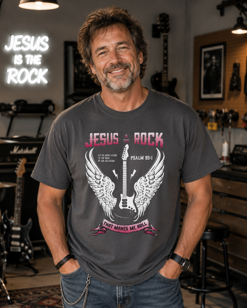 Man in his 50s with messy brown hair wearing a graphite Jesus Is the Rock Christian t-shirt in a modern garage music room with guitars, amps, and a clean contemporary studio atmosphere.