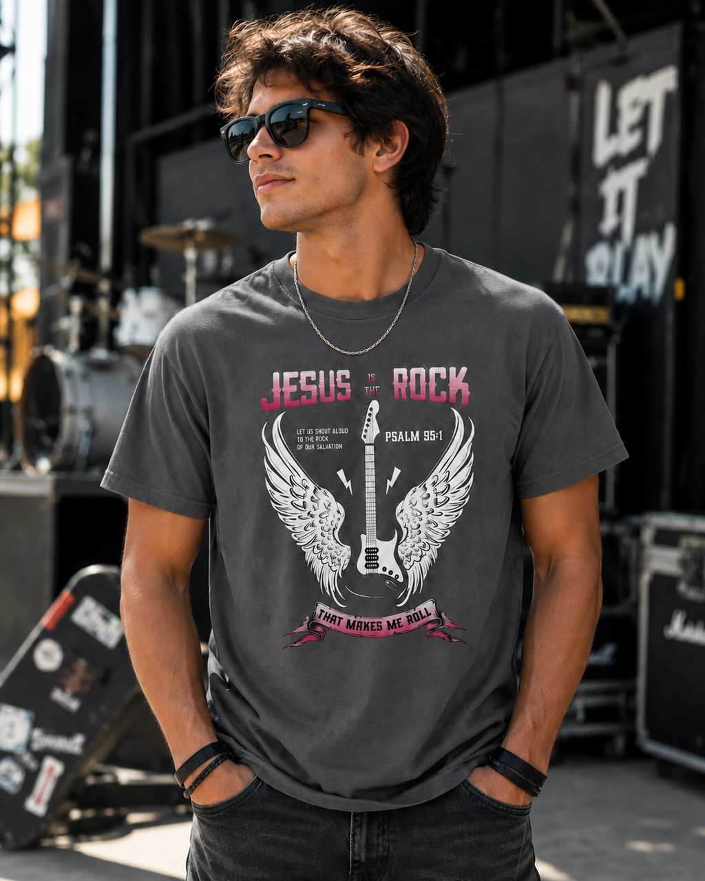 Young man in his 20s wearing a graphite Jesus Is the Rock Christian t-shirt at an outdoor concert venue with stage equipment, musical instruments, and a modern rock-inspired atmosphere.