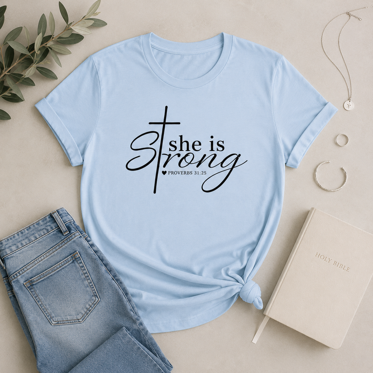 Light blue “She Is Strong” Proverbs 31:25 Christian women’s t-shirt flat lay with silver jewelry, denim, and neutral minimalist styling