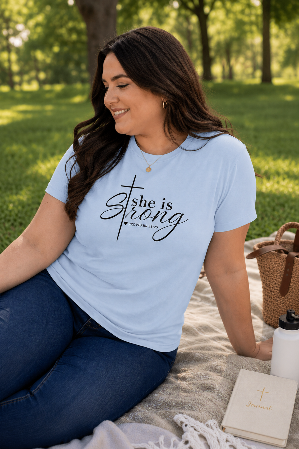 Plus-size brunette woman wearing light blue “She Is Strong” Proverbs 31:25 Christian t-shirt while sitting on a blanket in a sunny park setting