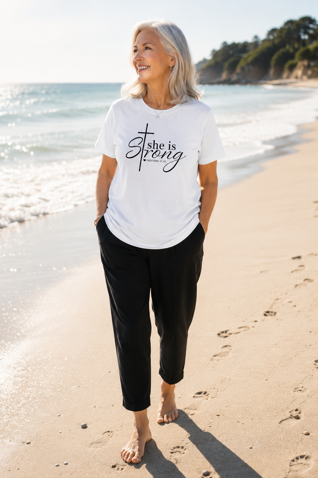 Older woman with shoulder-length gray hair wearing white “She Is Strong” Proverbs 31:25 Christian t-shirt and black pants while walking barefoot on a sunny beach