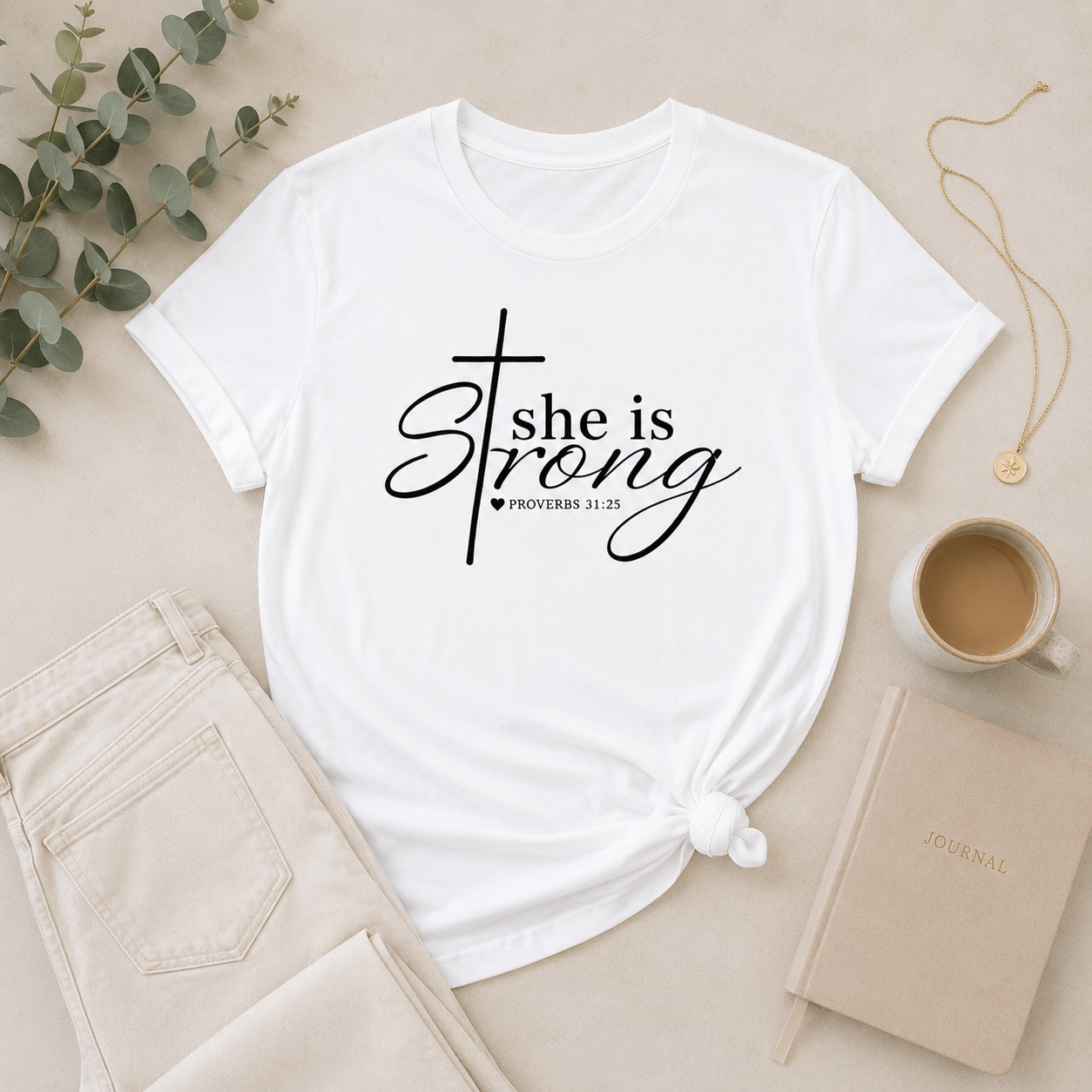 White “She Is Strong” Proverbs 31:25 Christian women’s t-shirt flat lay with neutral denim, gold jewelry, coffee, eucalyptus, and minimalist journal styling