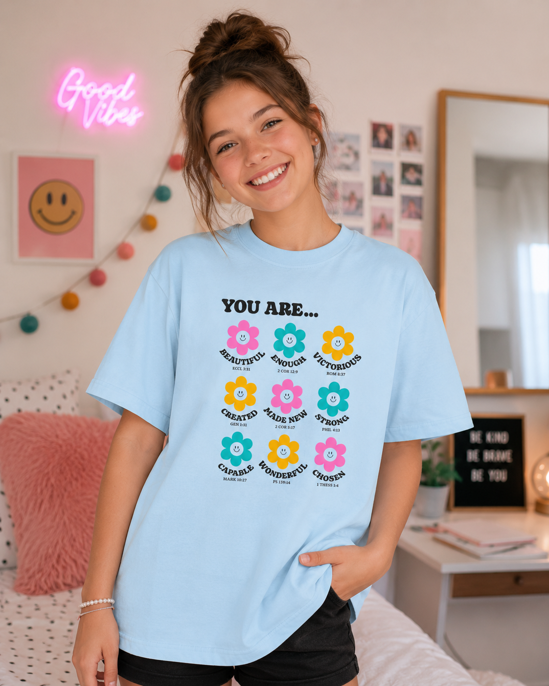 Tween brunette girl with a messy bun wearing a chambray blue “You Are…” Christian youth graphic t-shirt in a modern bedroom with colorful decor and a fun, uplifting aesthetic.