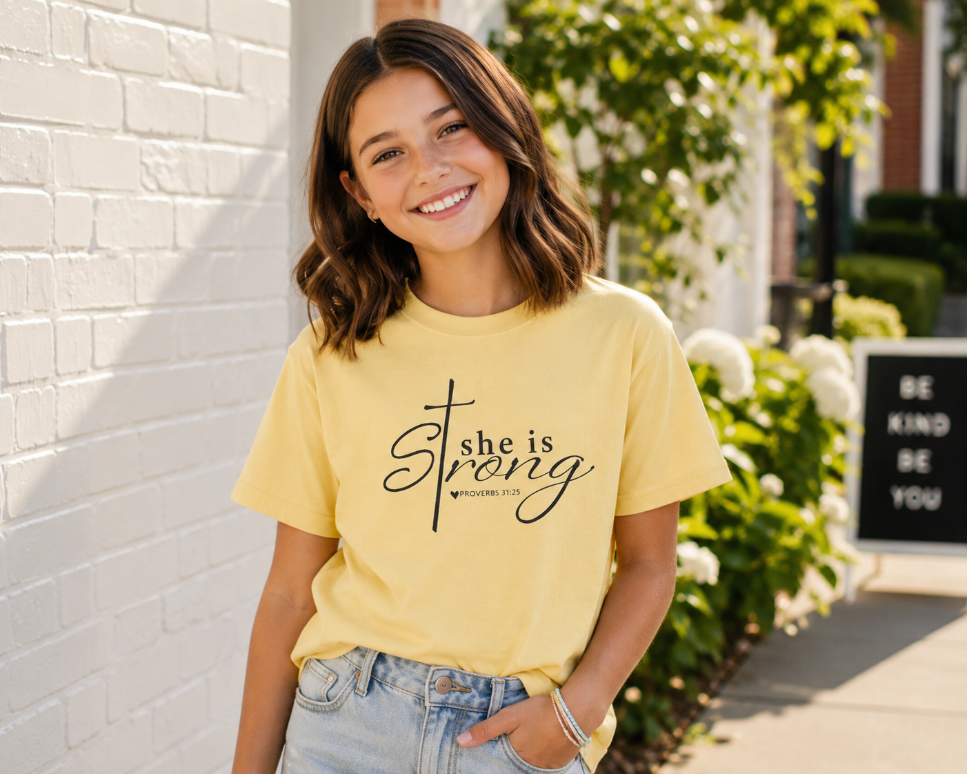 Tween girl with shoulder-length brunette hair wearing a butter yellow “She is Strong” Christian youth t-shirt outdoors in a bright modern setting.