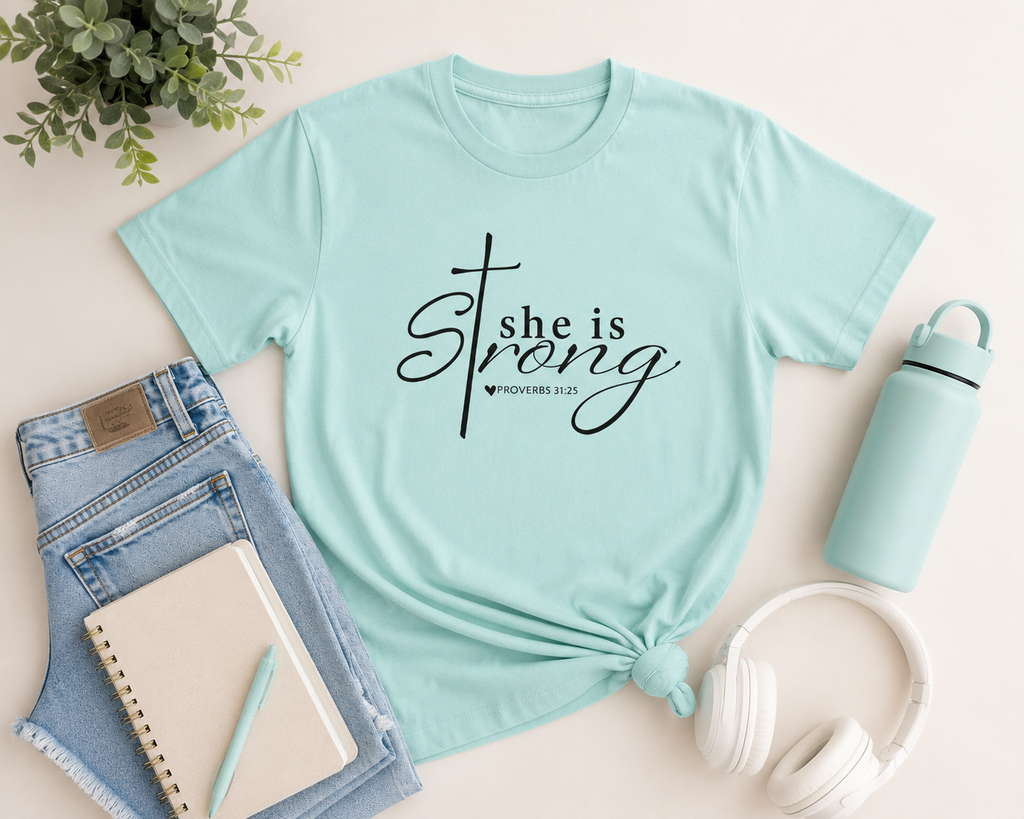 Flat lay mockup of the chalky mint “She is Strong” youth Christian t-shirt styled with denim shorts, wireless headphones, a notebook, and modern neutral accessories on a clean light background.