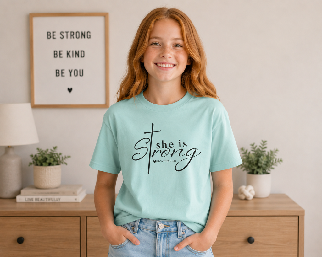Young strawberry blonde girl wearing the chalky mint “She is Strong” youth Christian t-shirt indoors with a modern dresser background and soft natural lighting.