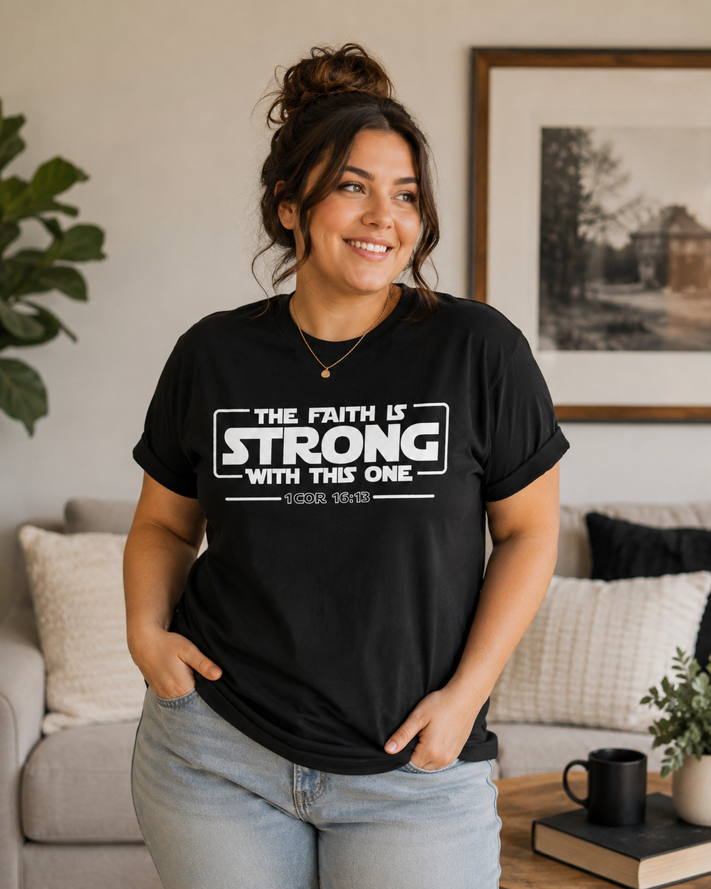 Plus-size brunette woman with a loose updo wearing a black Christian t-shirt with “The Faith Is Strong With This One 1 Cor 16:13” design while standing in a cozy modern living room with soft natural lighting