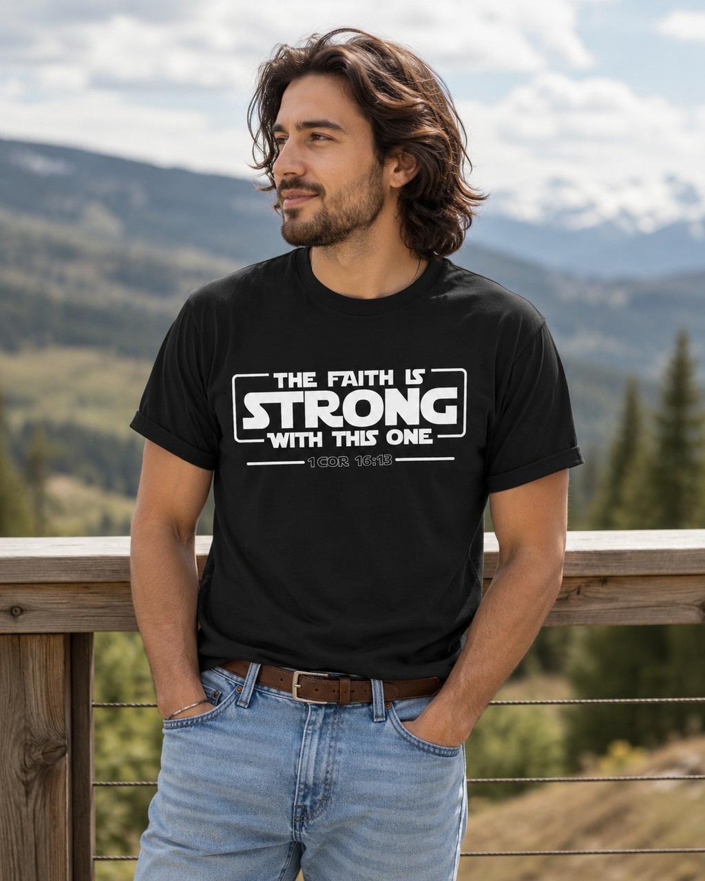 Young man wearing a black Christian t-shirt with “The Faith Is Strong With This One 1 Cor 16:13” design standing outdoors on a mountain deck railing, wearing jeans and looking to the side.