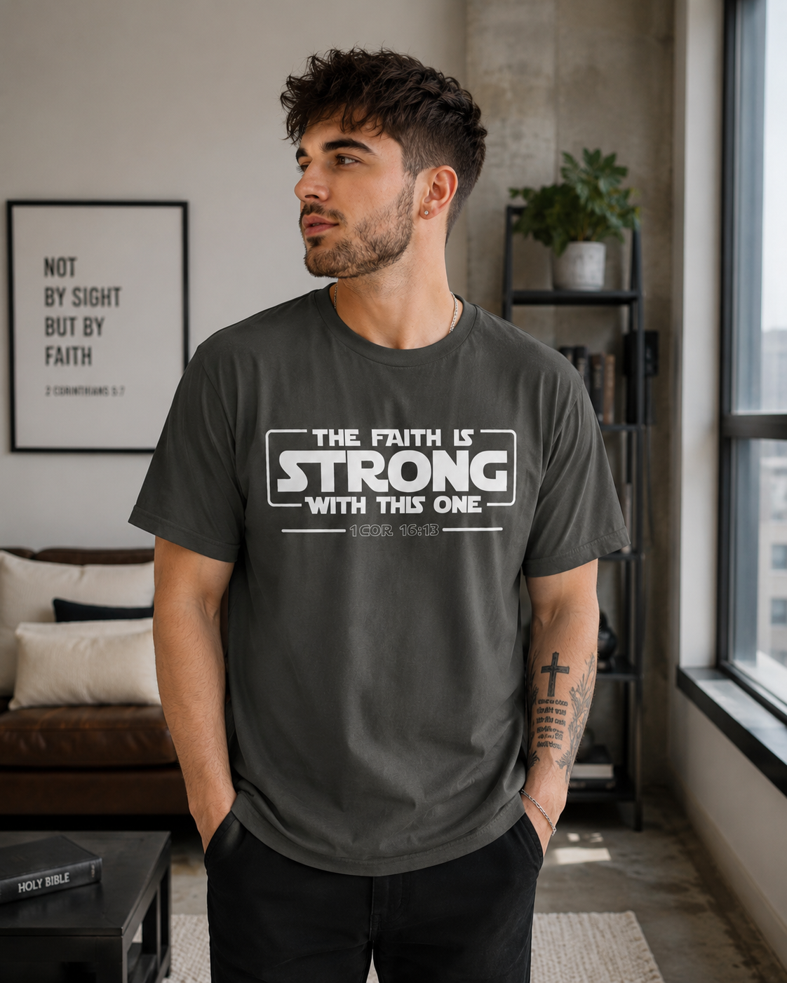 Young man with short dark hair and a Christian cross tattoo wearing a pepper-colored Christian t-shirt with “The Faith Is Strong With This One 1 Cor 16:13” design while standing in a modern industrial-style living room with soft natural lighting