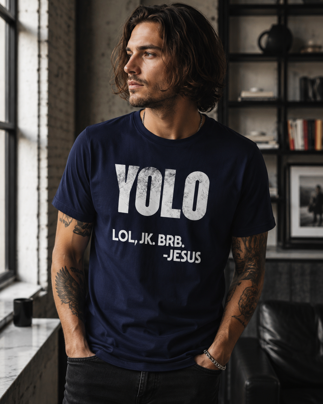 Man with chin-length hair wearing a true navy “YOLO LOL, JK. BRB. -Jesus” Christian graphic t-shirt while standing in a modern industrial-style interior with moody lighting and an edgy contemporary aesthetic