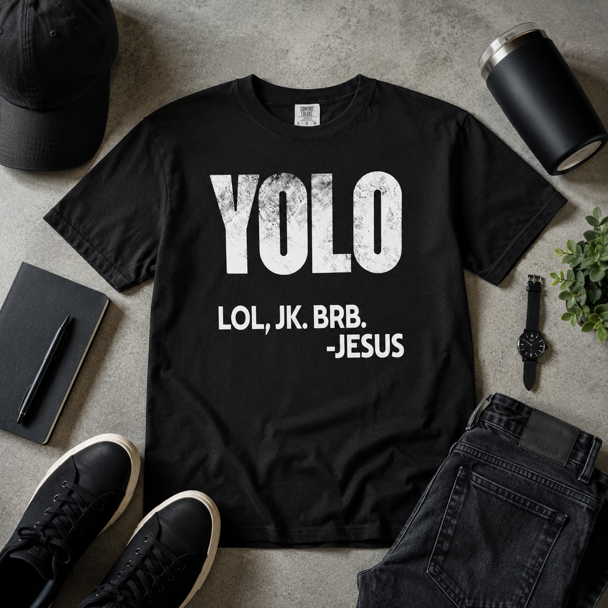 Black Christian humor t-shirt flat lay with distressed “YOLO LOL, JK. BRB. -Jesus” graphic styled with modern masculine accessories on a textured neutral background
