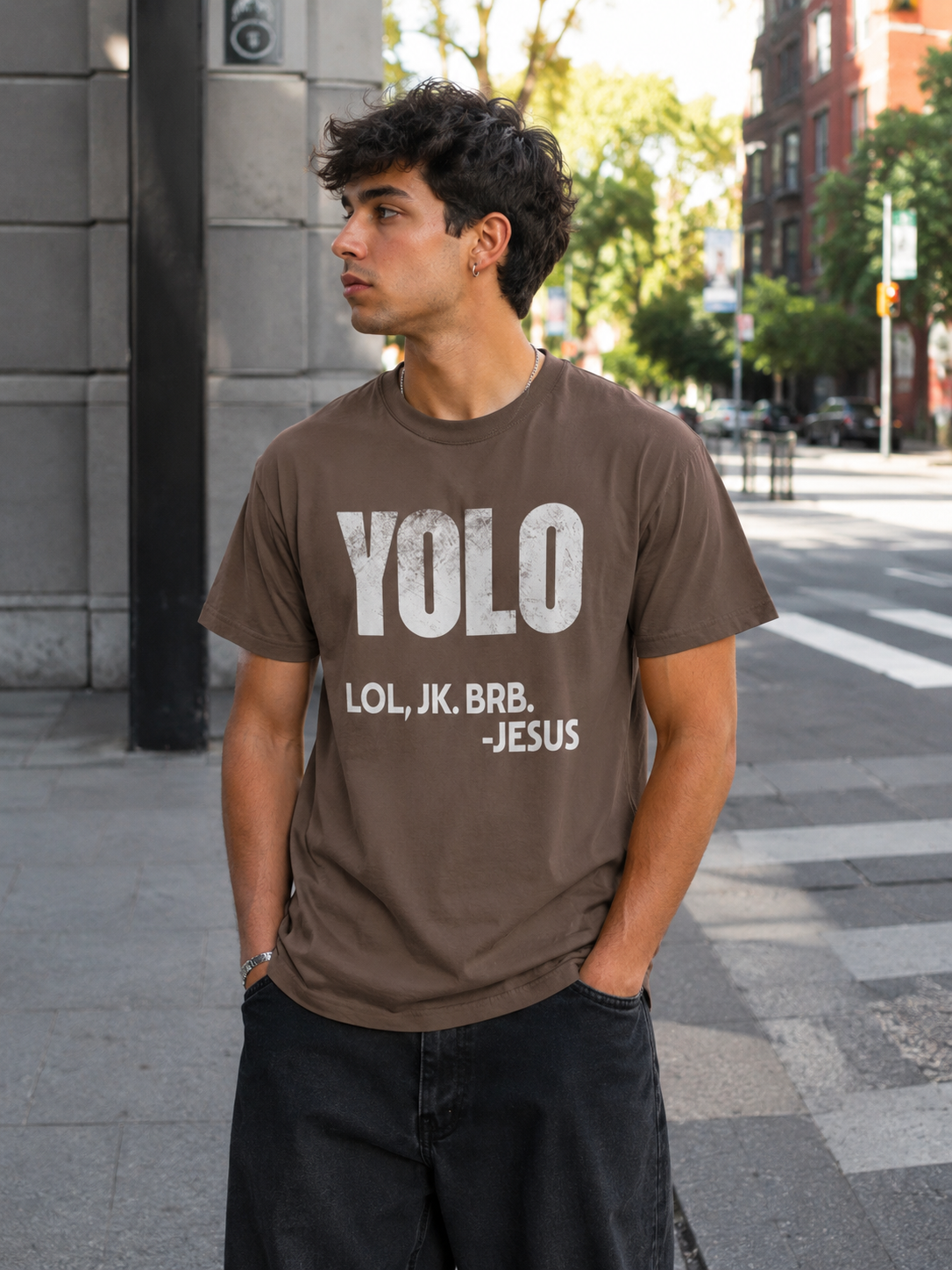 Young man wearing espresso brown Christian humor t-shirt with “YOLO LOL, JK. BRB. -Jesus” text while standing on a city street in dark relaxed-fit jeans