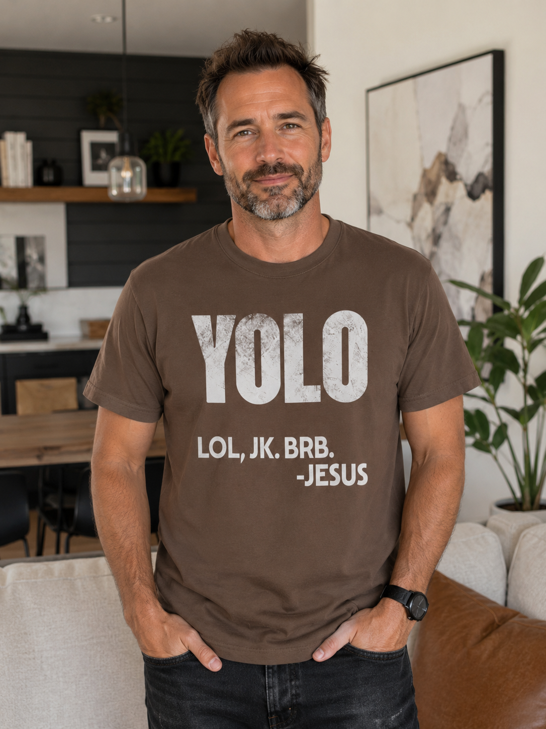 Middle-aged man wearing espresso brown Christian humor t-shirt with “YOLO LOL, JK. BRB. -Jesus” graphic while standing in a modern neutral-toned living room with soft natural light