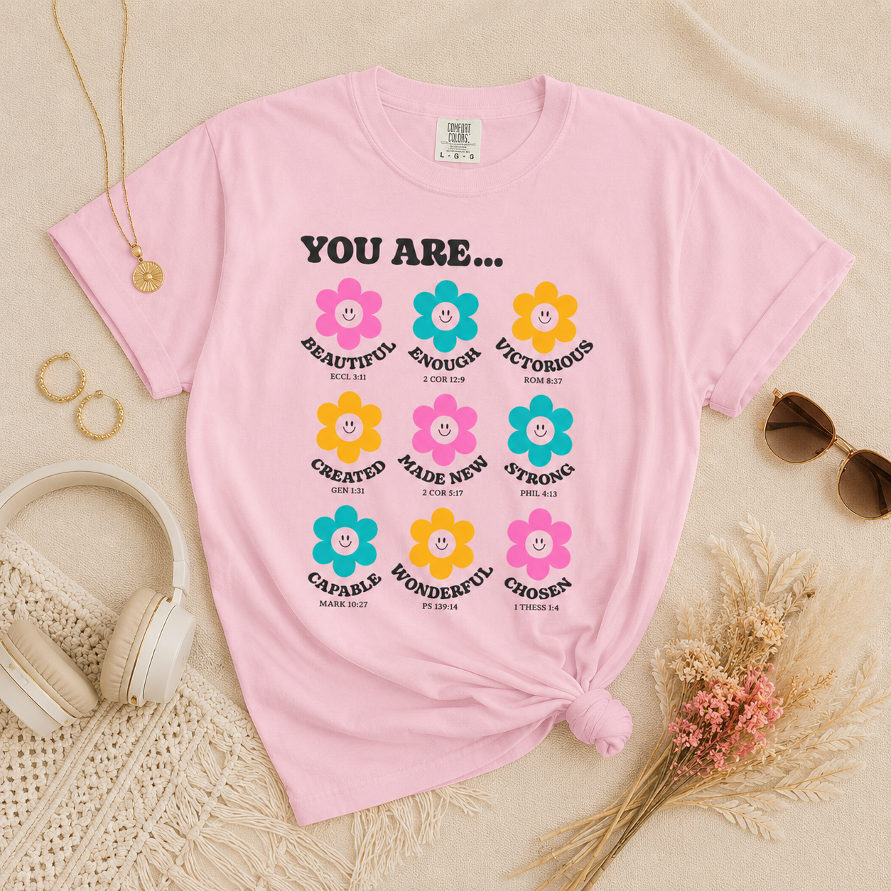 Blossom pink Christian affirmation flower t-shirt flat lay with cream headphones, gold jewelry, sunglasses, and dried florals on a soft neutral beige background