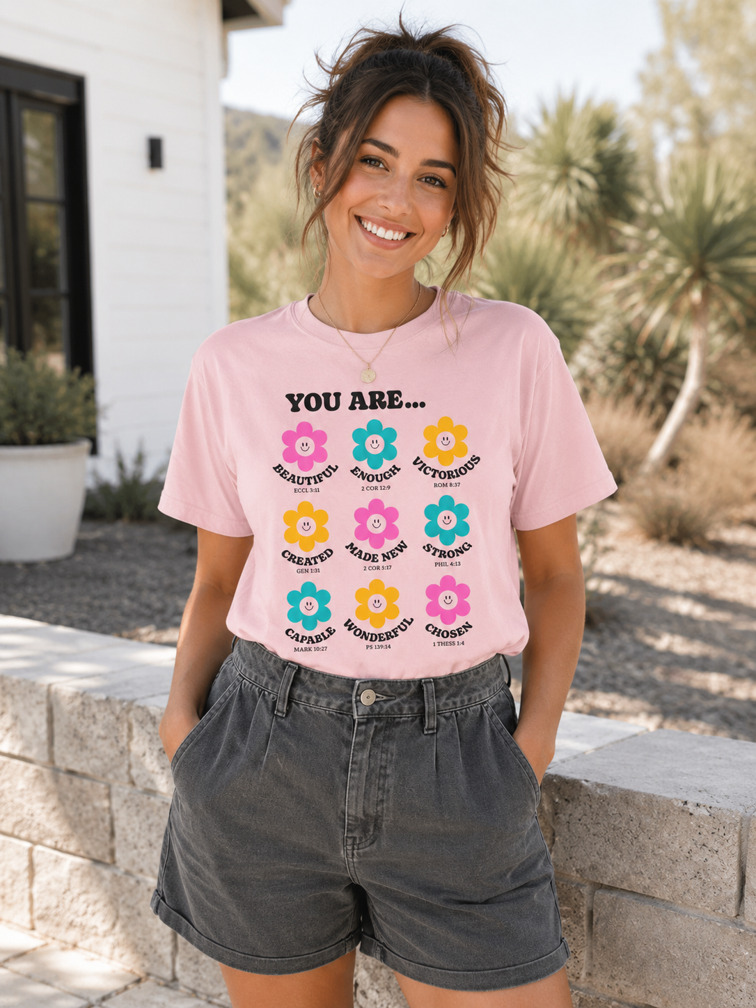 Young woman with messy ponytail wearing blossom pink Christian affirmation flower t-shirt tucked into charcoal shorts while smiling outdoors in soft natural light