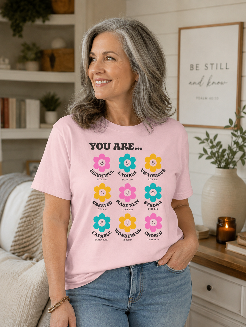 Older woman with shoulder-length salt and pepper hair wearing blossom pink Christian flower affirmation t-shirt in a cozy neutral-toned living room with soft natural lighting