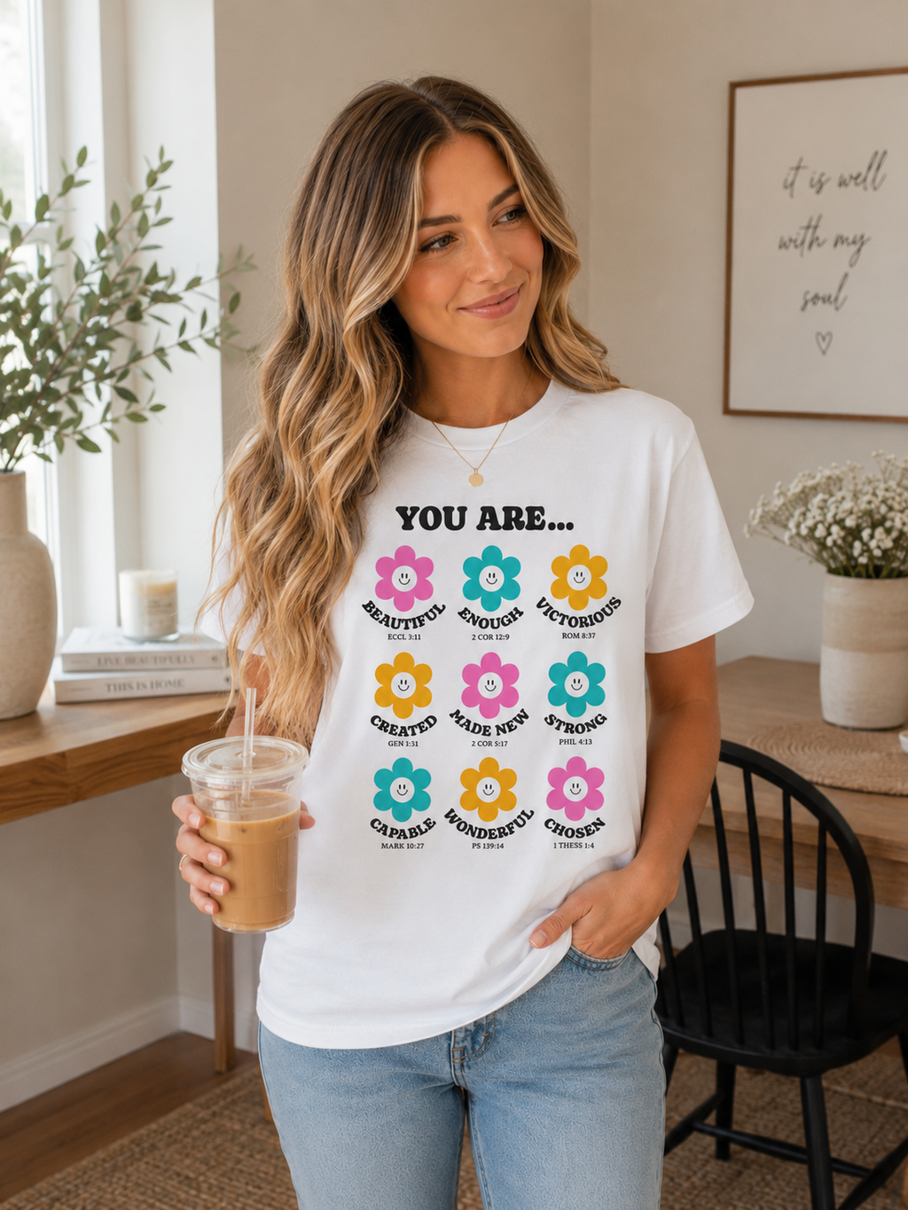Woman wearing white Christian flower affirmation t-shirt while holding iced coffee in a cozy neutral-toned kitchen and dining space with modern decor